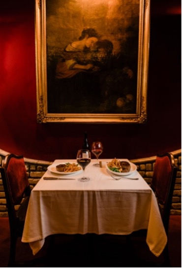 Fine dining table setting for two with a white tablecloth and two red-velvet chairs on either side, placed against red wall with framed painting above.